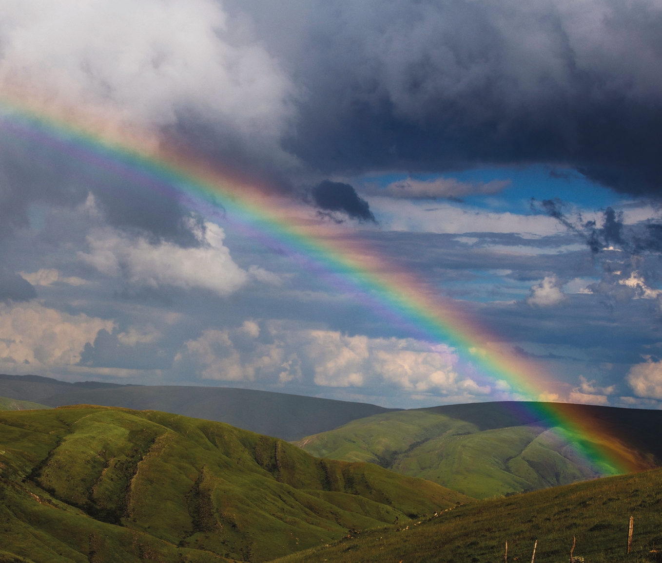 Fotografia. Um arco-íris atravessa o céu com nuvens escuras e claras até uma montanha coberta por vegetação.