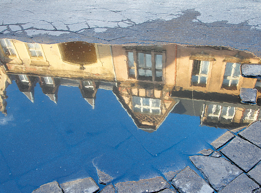 Fotografia. Uma construção de fachada amarela com várias janelas refletida em uma poça de água no chão