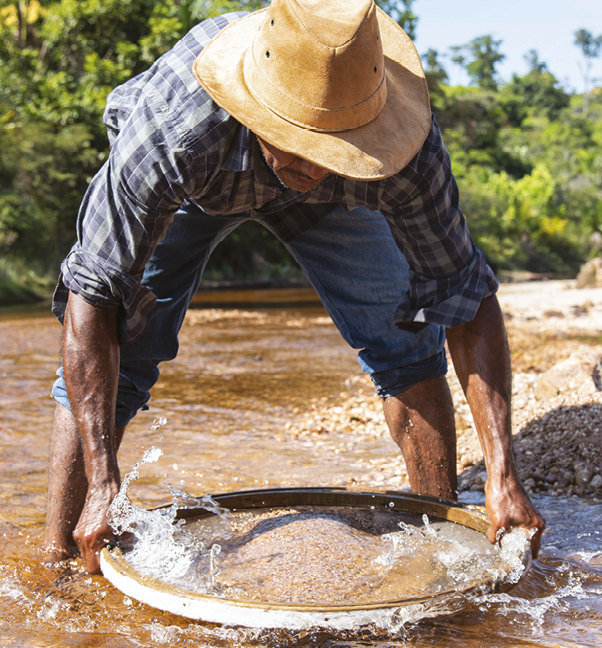 Fotografia. Um homem de calça jeans dobrada até o joelho, camisa azul xadrez e chapéu bege. Ele está em um rio, com a água até o calcanhar, com o tronco inclinado segurando uma peneira na superfície da água.