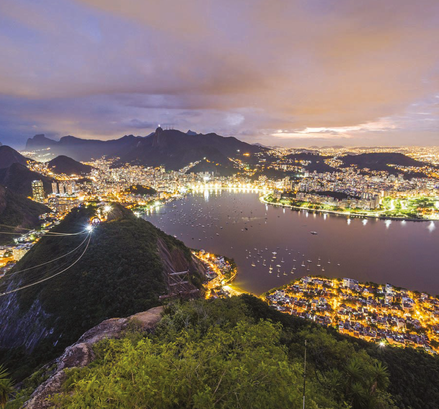 Fotografia. Vista aérea de parte de uma cidade durante a noite. As casas e prédios estão iluminadas artificialmente. No meio da cidade tem uma baia com barcos e em primeiro plano um morro coberto por vegetação.