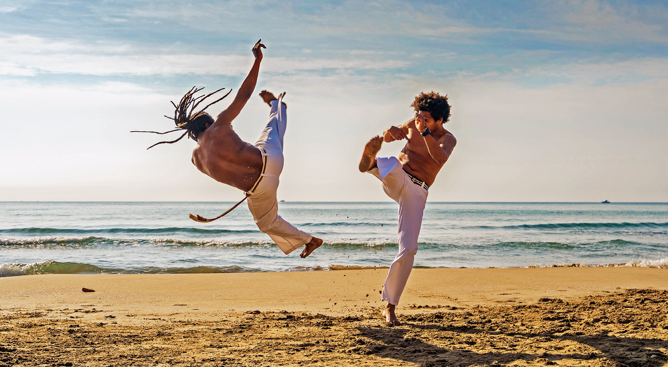 Fotografia. Dois homens em uma praia praticando capoeira. Ambos usam calça branca e cinto. Um deles tem o cabelo longo com tranças preso e está saltando com uma perna e um braço estendidos para cima. O outro tem o cabelo cacheado curto e está com uma perna levantada e os braços dobrados em frente ao corpo.