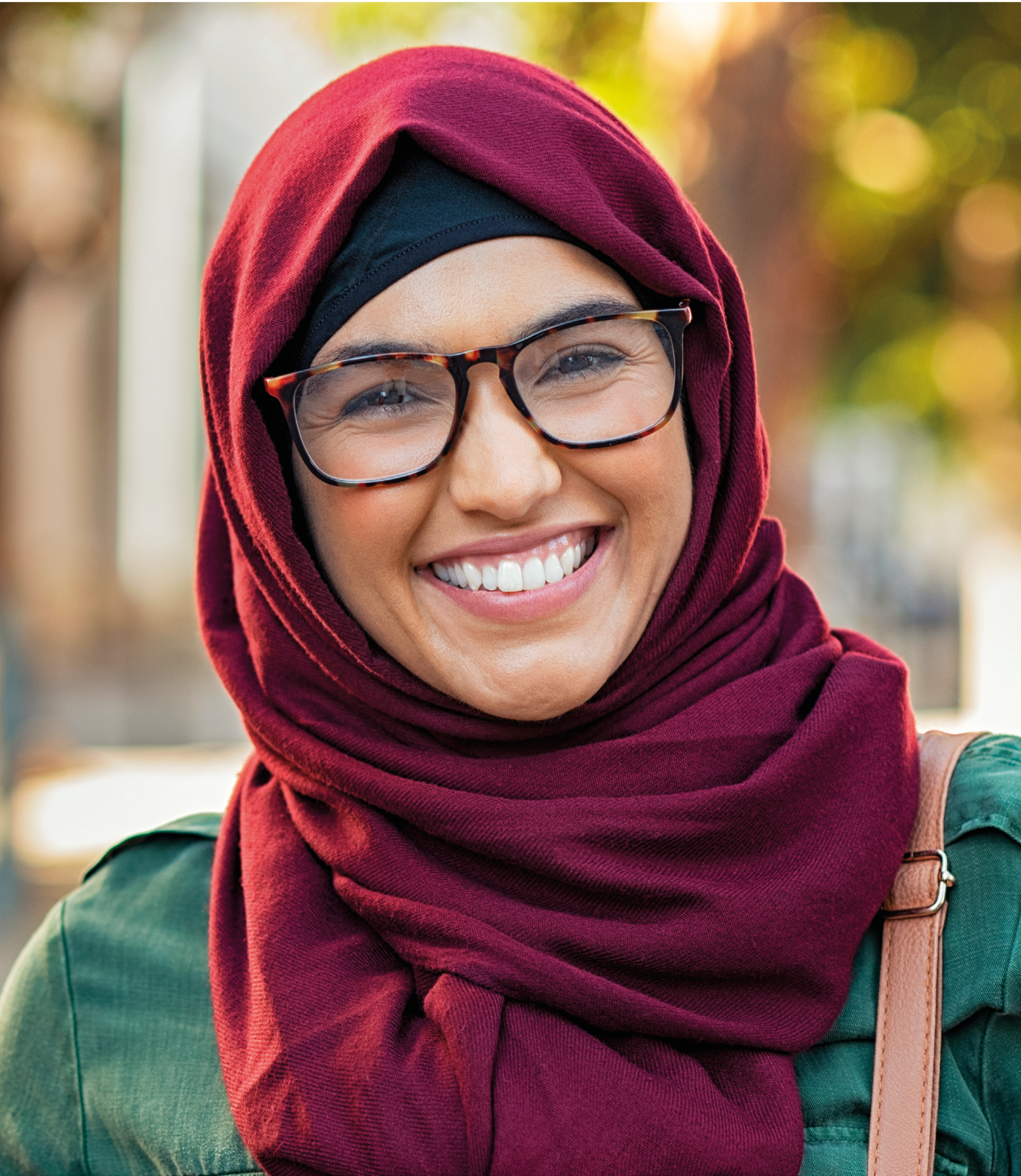 Fotografia. Mulher de óculos usando um hijabe roxo na cabeça e uma jaqueta jeans. Ela sorri.