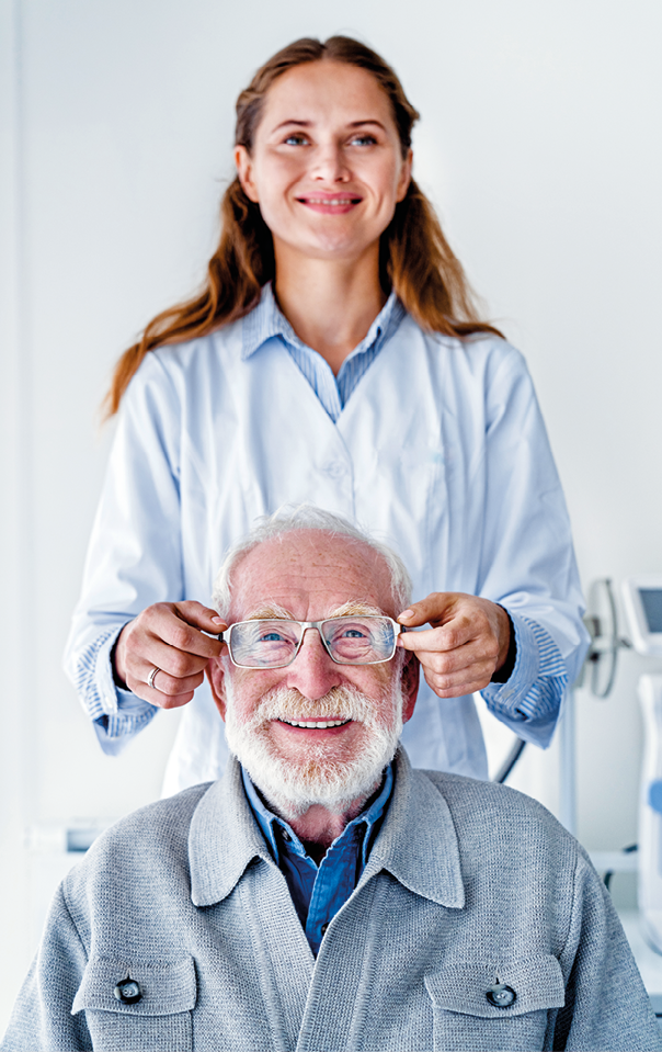 Fotografia. Mulher de cabelo castanho longo usando camisa azul e jaleco branco. Em pé, ela segura um par de óculos na frente do rosto de um homem idoso de barba vestindo suéter cinza, que está sentado na sua frente.