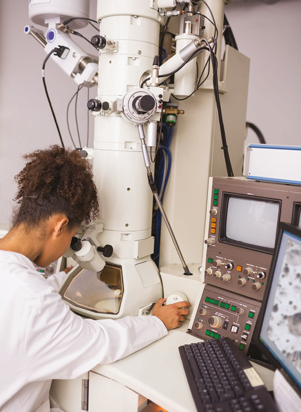 Fotografia. Mulher de cabelos castanhos e crespos presos em um rabo. Ela usa um jaleco branco e olha através de um microscópio eletrônico. Do lado, um computador.