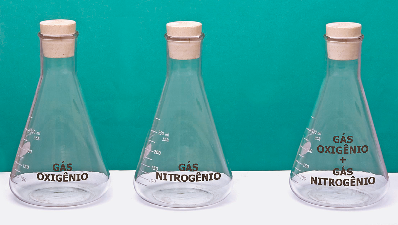 Fotografia. Três recipientes de vidro fechados com rolhas. No primeiro tem gás oxigênio, no segundo tem gás nitrogênio e no terceiro tem gás oxigênio + gás nitrogênio.