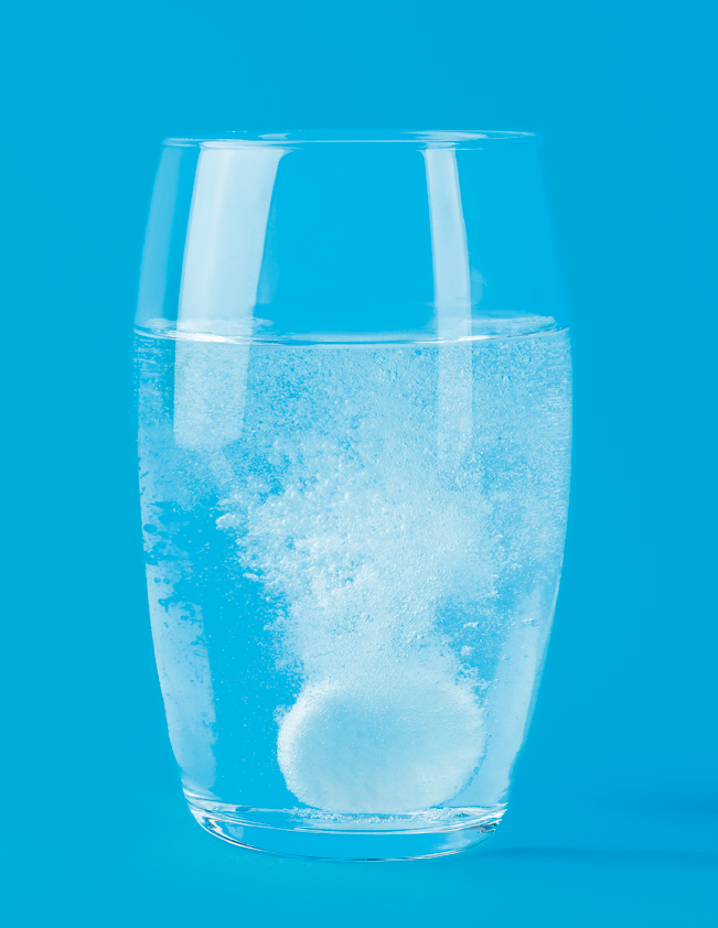 Fotografia. Um comprimido efervescente dentro de um copo de vidro com líquido transparente. Há várias bolhas saindo do comprimido.