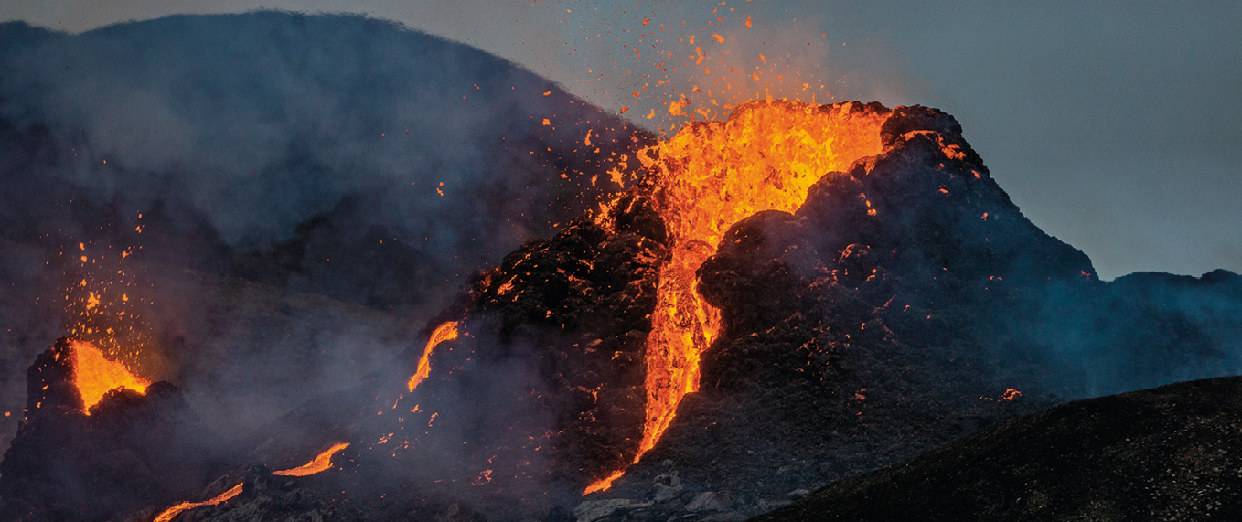 Fotografia. Um vulcão em erupção com lava de cor alaranjada saindo da parte superior e escorrendo pela superfície preta do vulcão.
