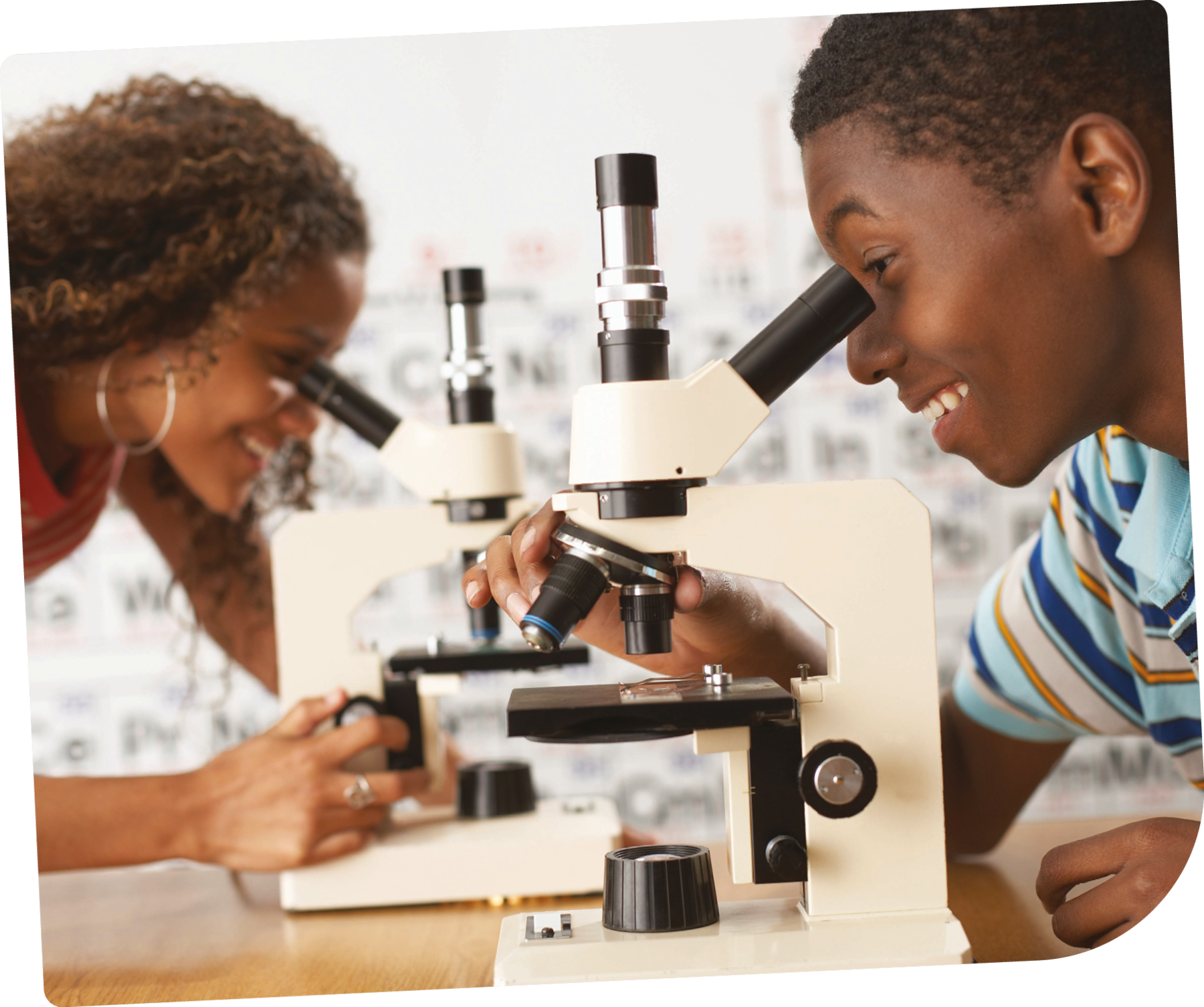 Fotografia. Dois adolescentes observando material em microscópios. Adolescente negra de cabelo cacheado, usa brincos de argola e camiseta vermelha. Adolescente negro de cabelo curto, usa uma camiseta azul com listras azuis escuras e laranjas.