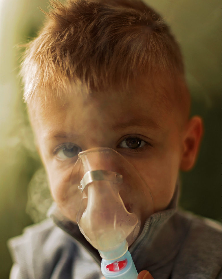 Fotografia. Criança loira de cabelo curto com um inalador soltando fumaça na frente do rosto.