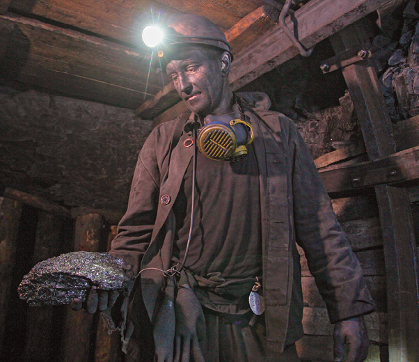 Fotografia. Homem dentro de uma mina, com paredes escoradas por estacas de madeira. Ele usa um capacete com uma lanterna acesa, roupa marrom e segura um fragmento de rocha nas mãos. O rosto dele está sujo de terra e ele tem uma máscara pendurada no pescoço.