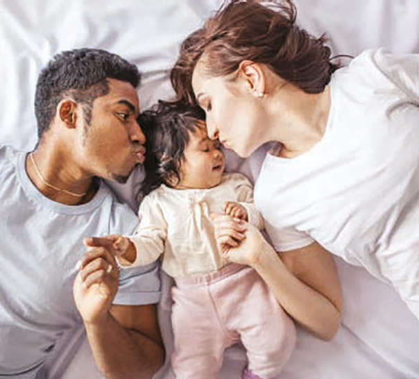 Fotografia. Um homem e uma mulher deitados na cama dando um beijo em uma bebê que está deitada entre eles. Cada um segura uma mão da bebê. O homem é negro e tem o cabelo preto crespo e curto, usa uma camiseta cinza, e a mulher é branca e tem o cabelo castanho e liso, veste uma camiseta branca. A bebê é morena e tem o cabelo preto liso, veste um macacão rosa.