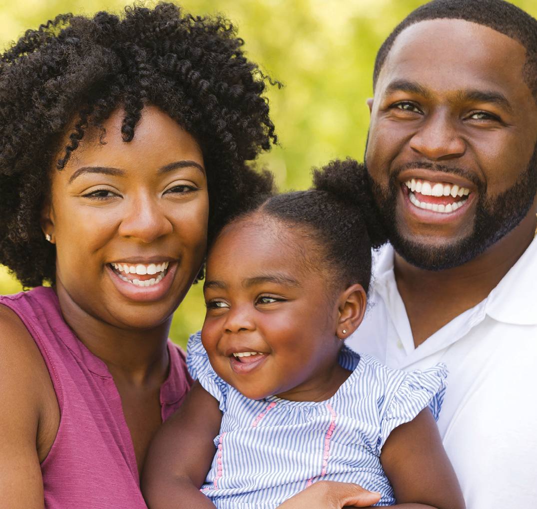 Fotografia. Homem e mulher com uma menina no colo. Os três sorriem. A mulher é negra, tem o cabelo preto crespo e usa uma camiseta regata rosa. O homem é negro com o cabelo preto bem curto, barba aparada e veste uma camisa branca. A menina é negra, tem o cabelo preto e crespo, o qual está preso em maria-chiquinha; ela veste uma camiseta azul listrada.