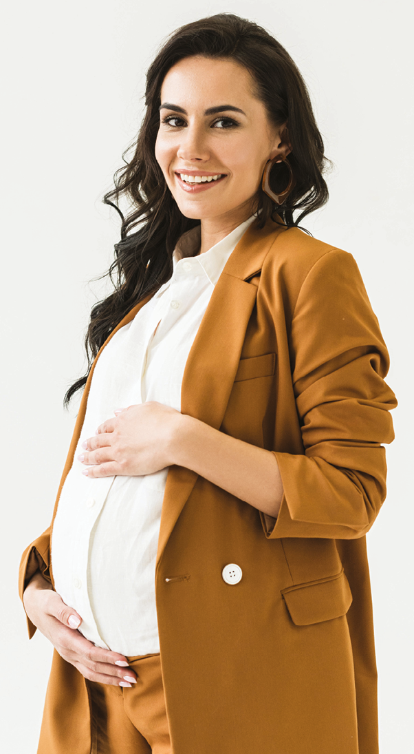 Fotografia. Mulher grávida com as mãos na barriga. Ela tem cabelo preto comprido, usa camiseta branca e casaco marrom.
