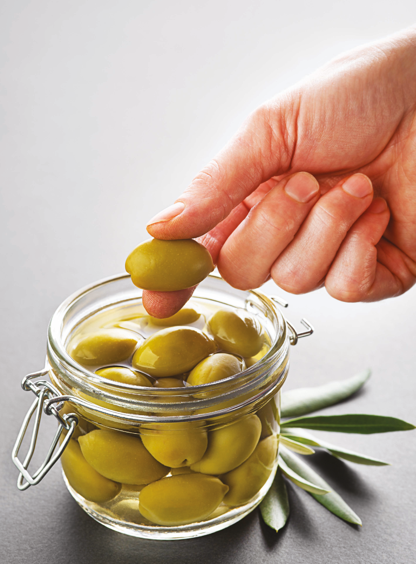 Fotografia. Destaque para a mão de uma pessoa segurando uma azeitona verde. Embaixo, pote de vidro com mais azeitonas em um líquido. Embaixo do pote há algumas folhas de oliveira.