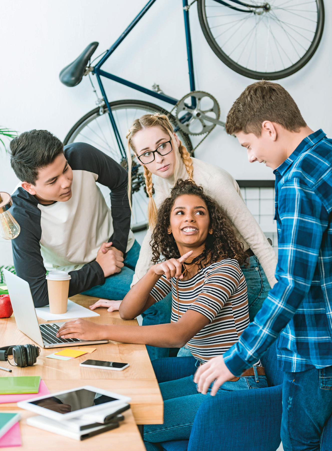 Fotografia. Quatro jovens ao redor de uma mesa. Na mesa tem computador, celular, tablet, fone de ouvido, um copo e papéis. Na frente do computador, menina morena de cabelo longo cacheado e camiseta listrada. Ela aponta para um menino branco de cabelo castanho e camisa xadrez azul. Ao lado deles, menina branca com o cabelo loiro preso em duas tranças, ela usa óculos e uma camiseta de gola alta branca. Do lado dela, apoiado na mesa, menino asiático de cabelo preto liso e camiseta branca com manga longa preta.