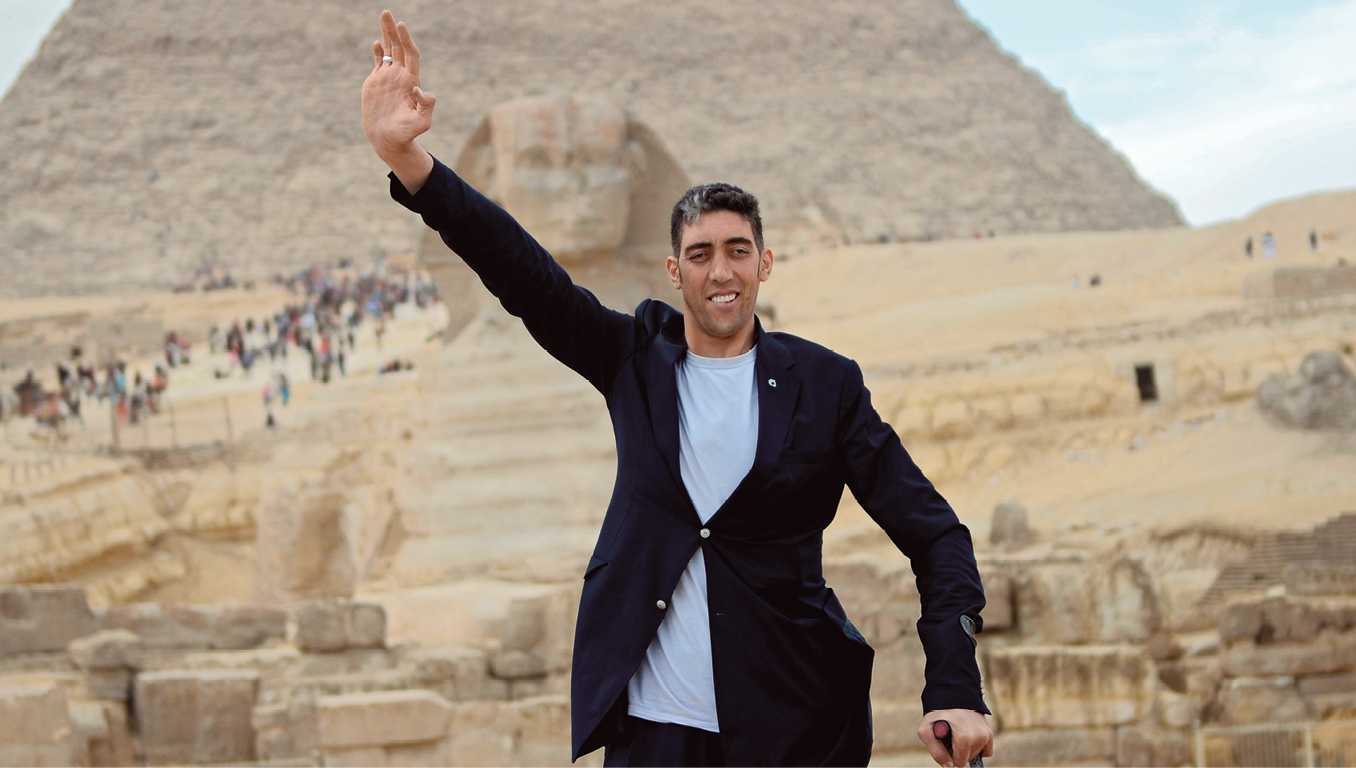 Fotografia. Homem muito alto, de cabelo preto usando uma camiseta branca e um terno azul escuro. Ele está no Egito, em frente a uma pirâmide com um braço erguido.