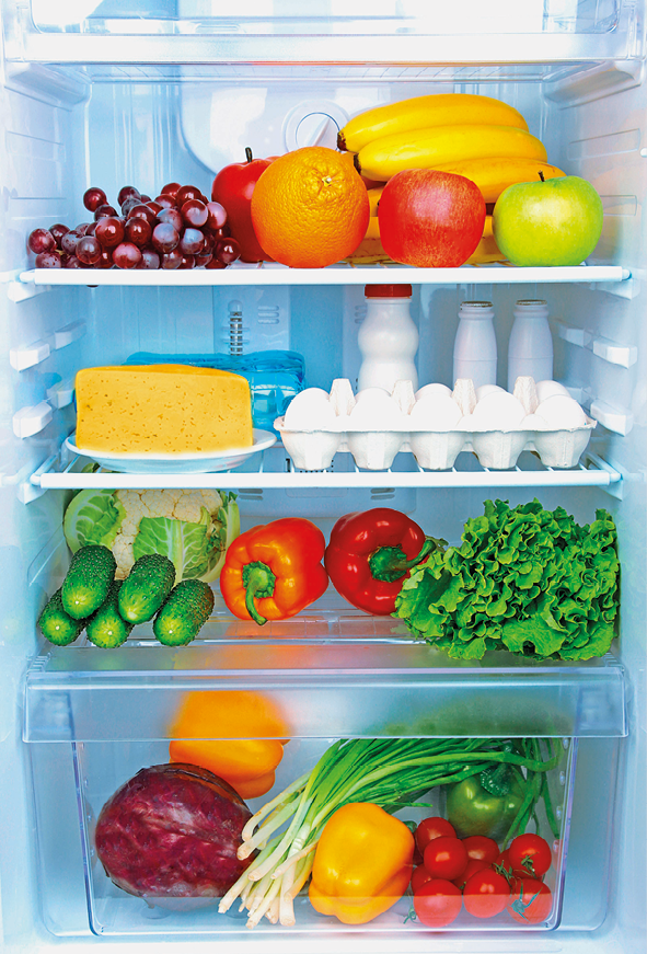 Fotografia. Parte interna de uma geladeira. Dentro, nas prateleiras há frutas (banana, maçã, uva, laranja), um prato com queijo, ovos, leite, pepino, pimentão, alface, repolho, cebolinha e tomate.
