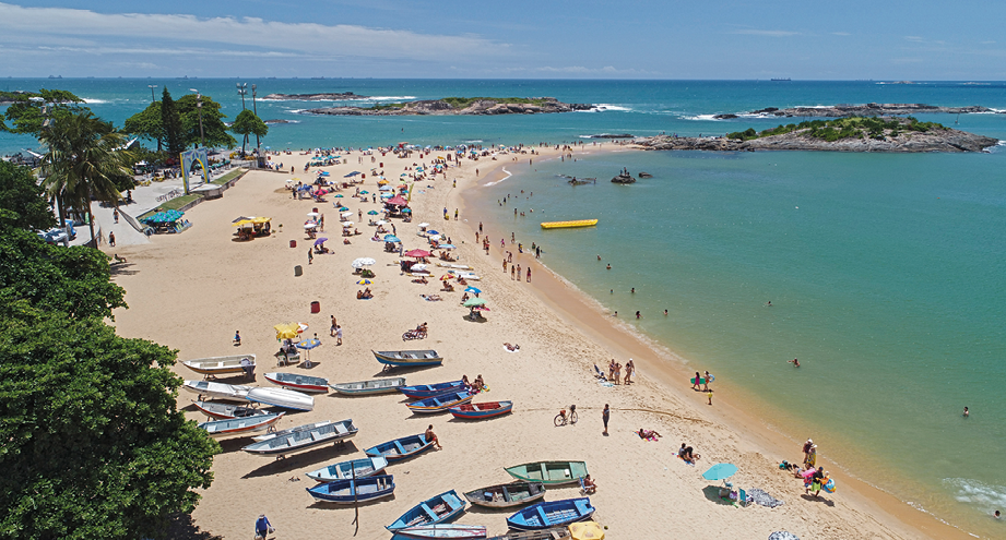 Fotografia. Praia com pessoas caminhando na margem da água, nadando ou sob guarda-sóis. Ao lado, canoas coloridas na areia. Atrás, uma faixa de coqueiros.