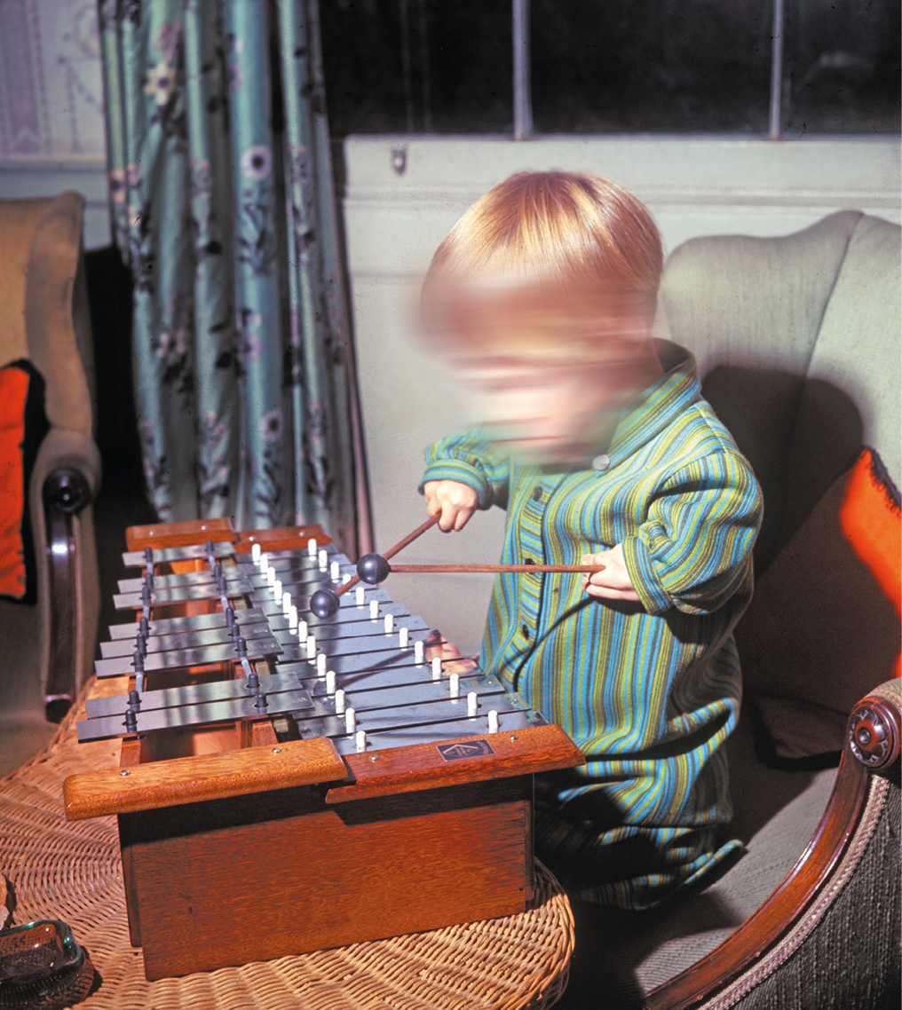 Fotografia. Criança branca, com cabelos loiros e lisos. Seus braços são curtos, próximo ao corpo. Ela está tocando um xilofone.