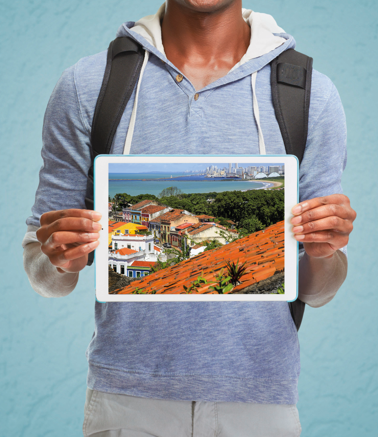 Fotografia. Rapaz de moletom cinza e mochila preta nas costas. Ele segura um tablet na frente do corpo. Na tela do tablete, imagem de uma cidade com sobrados coloridos e a praia ao fundo.