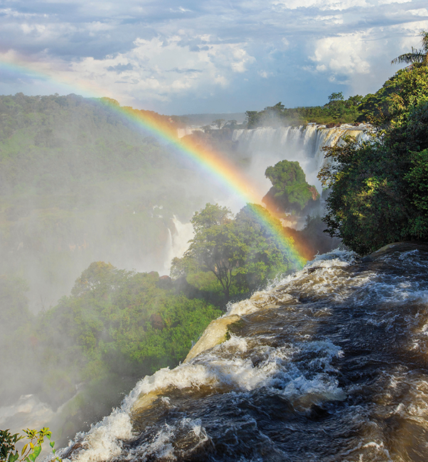 Fotografia. Cachoeira no meio de uma floresta. Um arco-íris corta o céu.