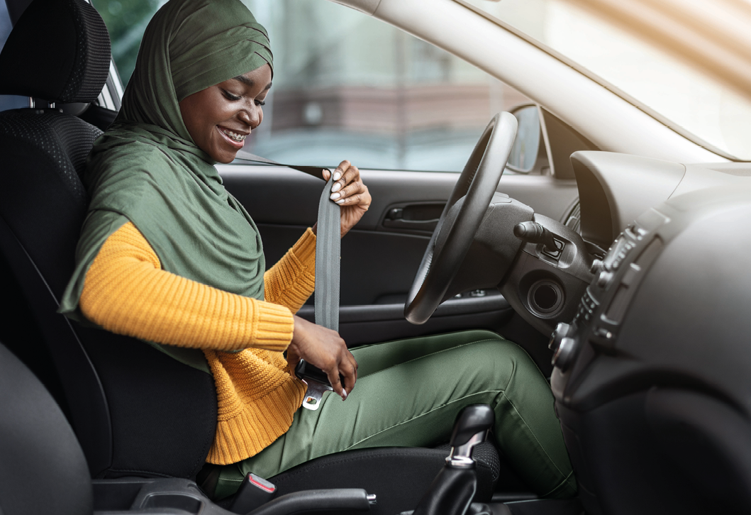 Fotografia. Mulher negra com um lenço verde na cabeça e blusa amarela. Ela está sentada no banco do motorista de um carro e coloca o cinto de segurança.