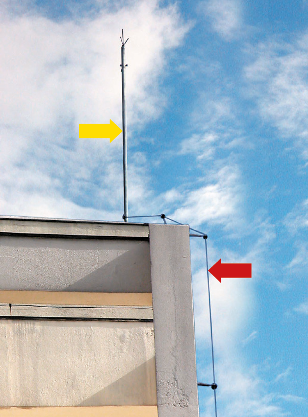 Fotografia. Telhado de uma edificação com uma haste metálica vertical, indicado por uma seta amarela, e um fio que desce paralelo à parede do prédio, indicado por uma seta vermelha.
