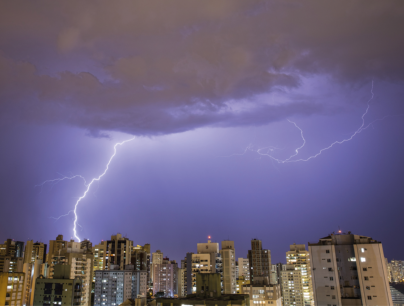 Fotografia. Parte de um centro urbano, com diversos prédios, durante a noite. Dois relâmpagos saindo de nuvens iluminam o céu noturno.