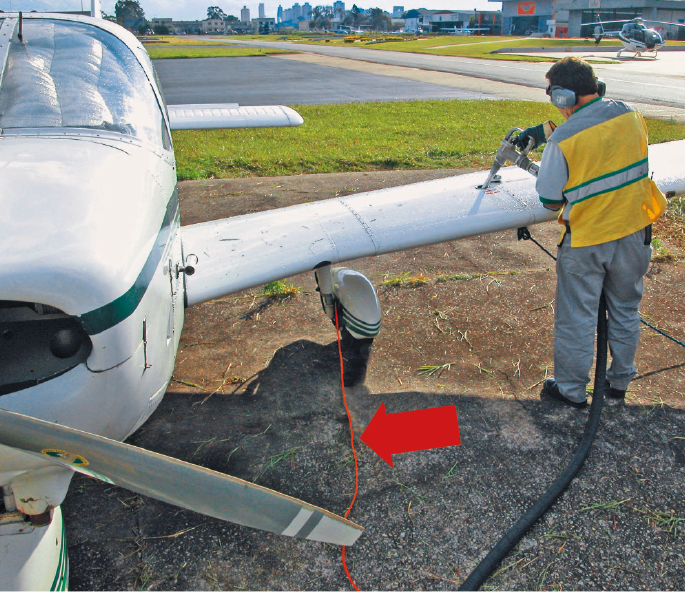 Fotografia. Uma pessoa com colete de proteção amarelo e abafador nos ouvidos. Ela coloca combustível na asa de um avião. Da roda do avião sai um fio vermelho, indicado por uma seta vermelha.