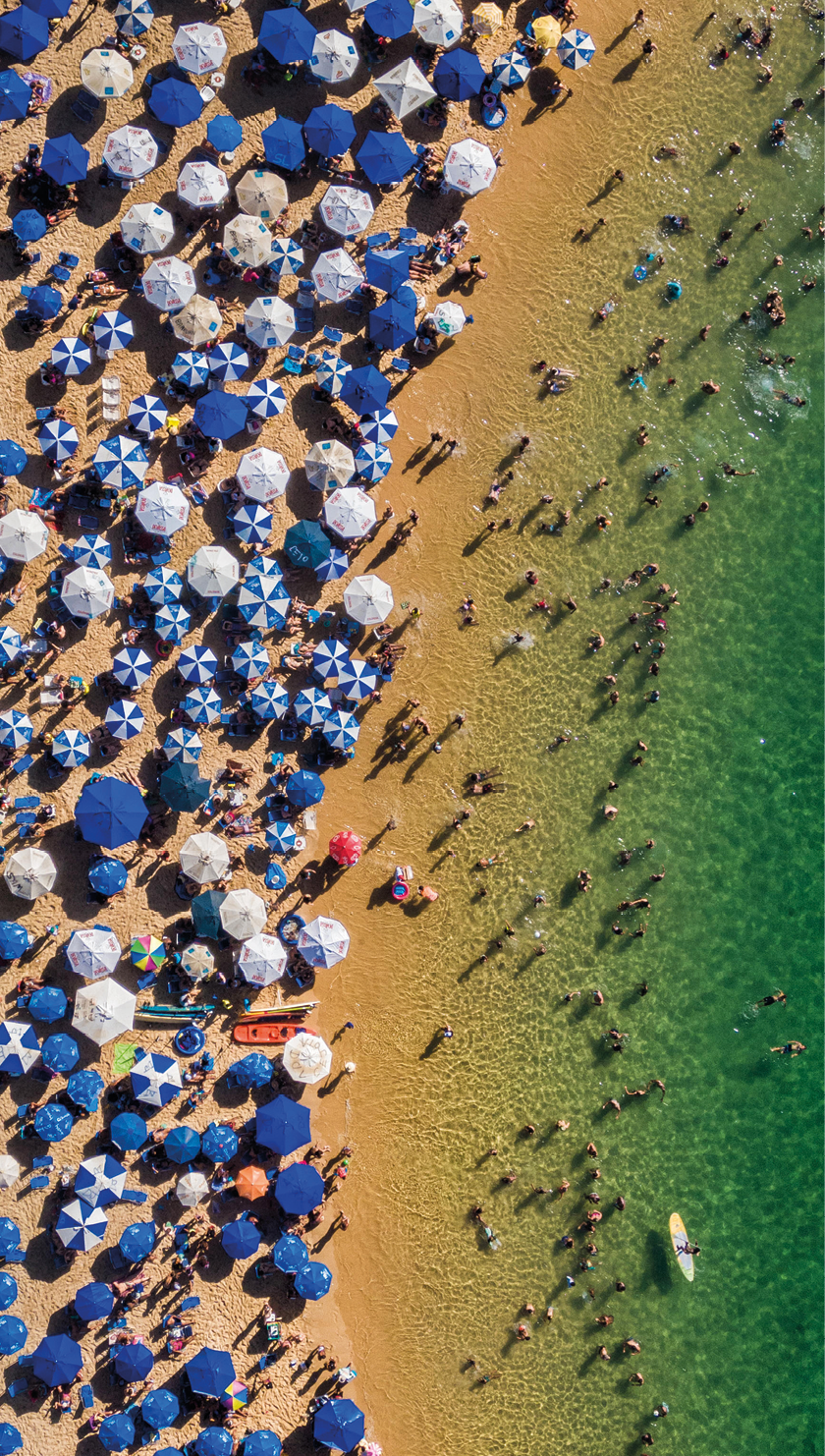 Fotografia. Imagem aérea de uma praia com pessoas no mar e guarda-sóis azuis e brancos na areia.