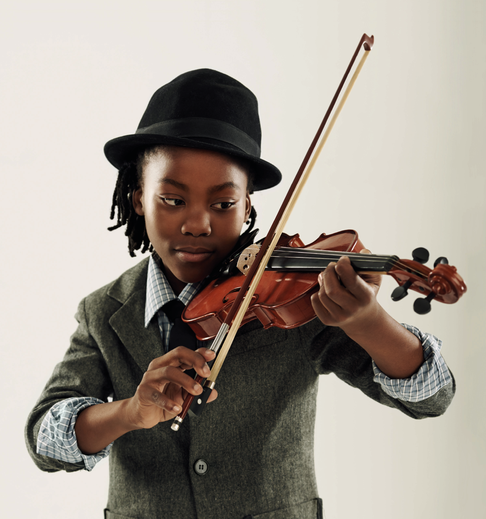 Fotografia. Menino negro de cabelo com tranças, veste uma camisa xadrez, terno e chapéu preto. O menino toca um violino.