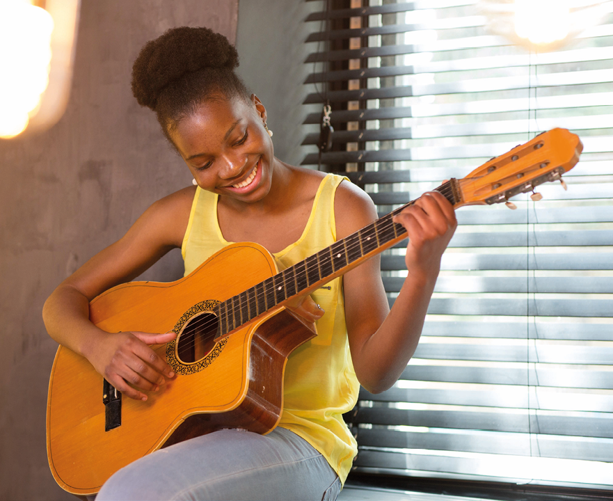 Fotografia. Mulher negra com o cabelo preto preso em um coque. Ela usa uma blusa regata amarela e está sentada tocando um violão.