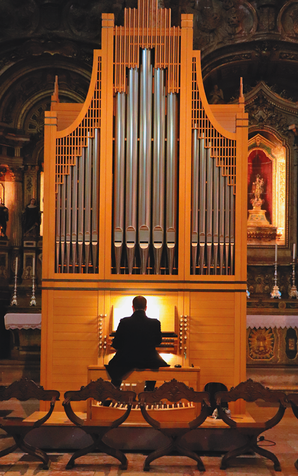 Fotografia. Homem sentado de costas em uma igreja. Ele está tocando um órgão alto formado por vários cilindros prateados na vertical, com diferentes alturas.