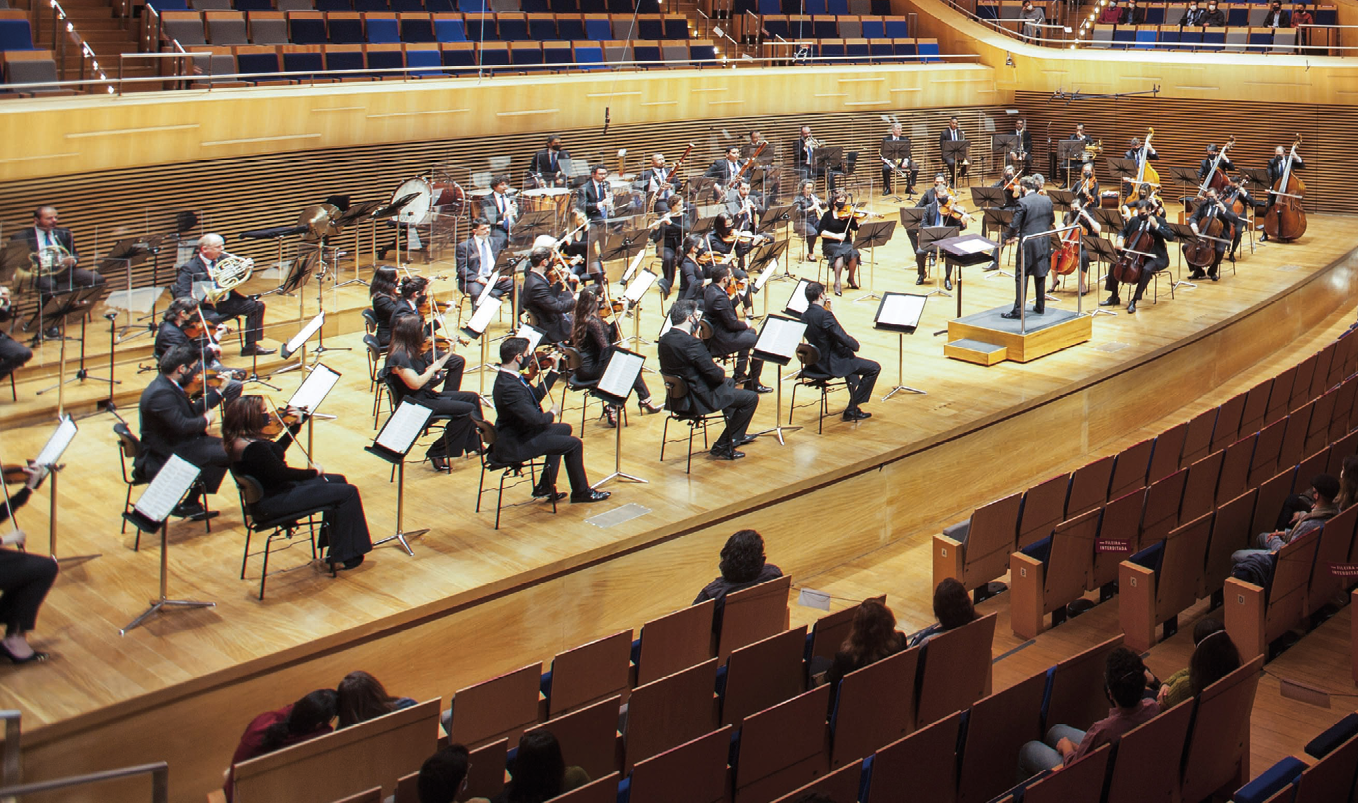 Fotografia. Uma orquestra em cima de um palco. As pessoas tocam diversos instrumentos e no centro tem um maestro. Na frente, plateia com algumas pessoas.