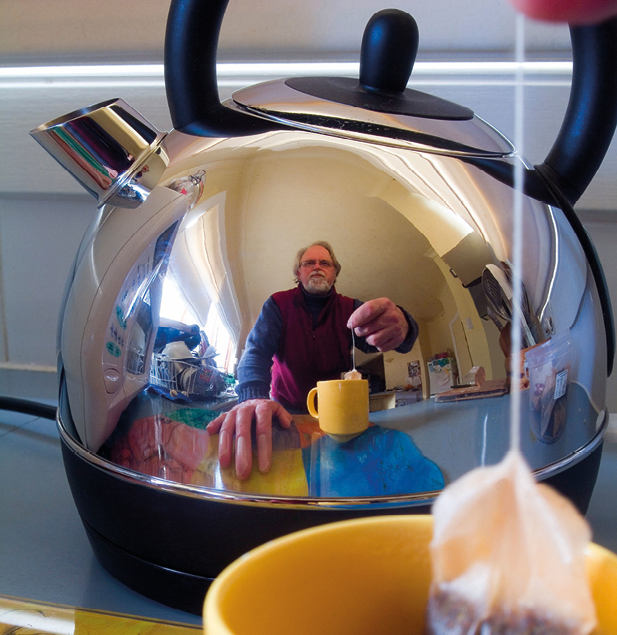 Fotografia. Reflexo de um homem colocando um sachê de chá em uma xícara amarela. O reflexo está na superfície de uma chaleira de inox.