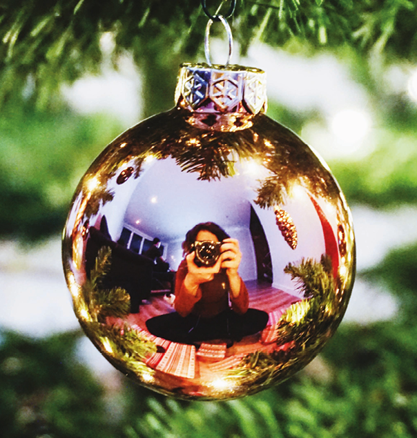 Fotografia. Reflexo de uma pessoa sentada de pernas cruzadas, segurando uma máquina fotográfica. O reflexo está em uma bolinha espelhada de decoração de Natal.