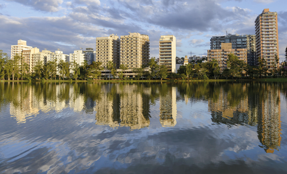 Fotografia. Vista de um lago refletindo parte de uma cidade, com prédios e árvores. A imagem não é refletida perfeitamente.