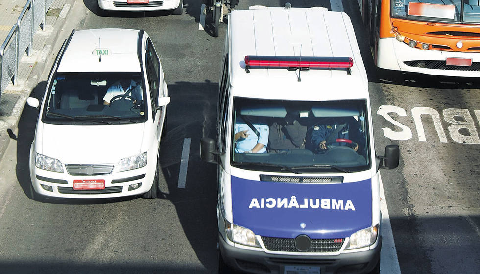 Fotografia. Carros em uma via pública. No meio, uma ambulância com a palavra “ambulância” no capô azul, escrita da direita para a esquerda.