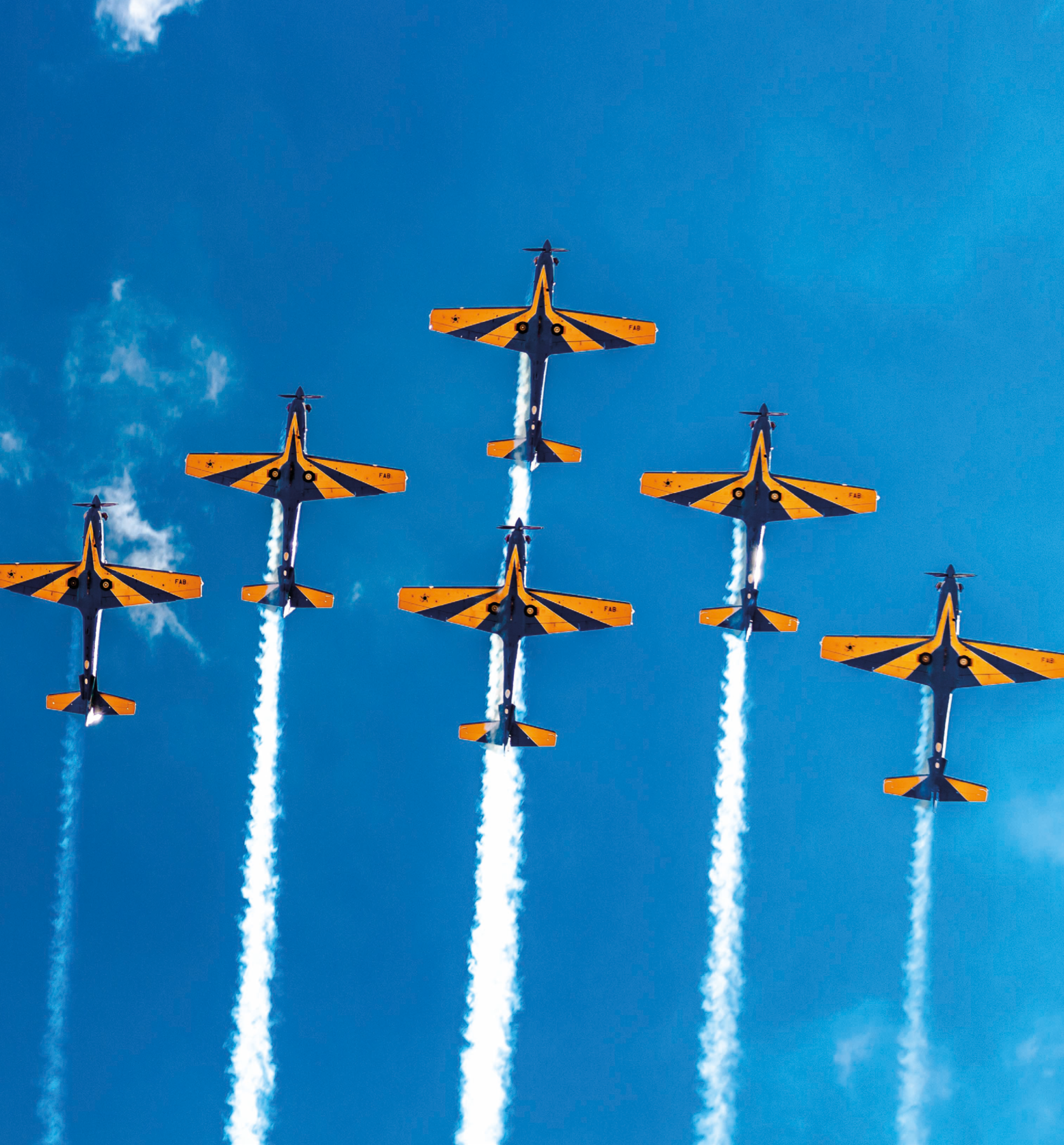 Fotografia. Seis aviões da força aérea no céu, voando formando um "V" no céu. Eles são amarelos e azuis e há uma faixa de fumaça atrás de cada um.