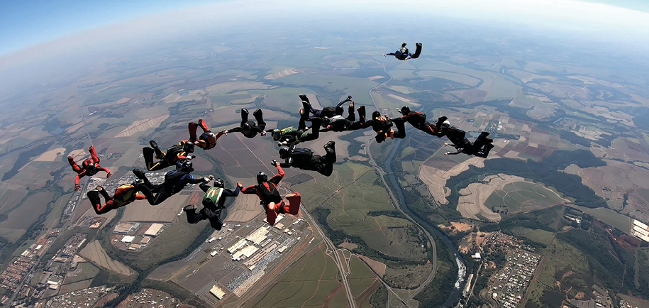 Fotografia. Um grupo de pessoas saltando de paraquedas. Eles estão de mãos dadas formando um círculo no céu.