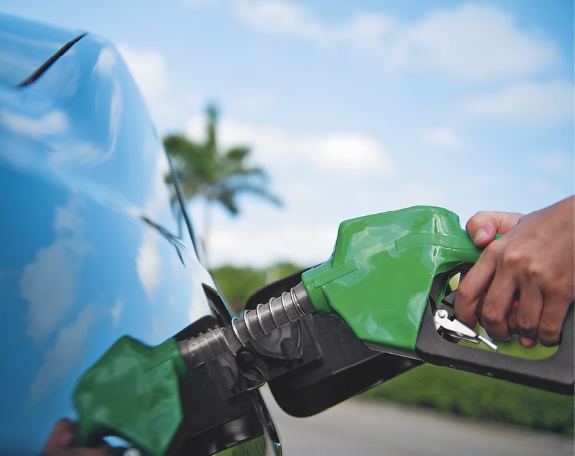Fotografia. Destaque para a mão de uma pessoa segurando uma mangueira no tanque de combustível de um automóvel.