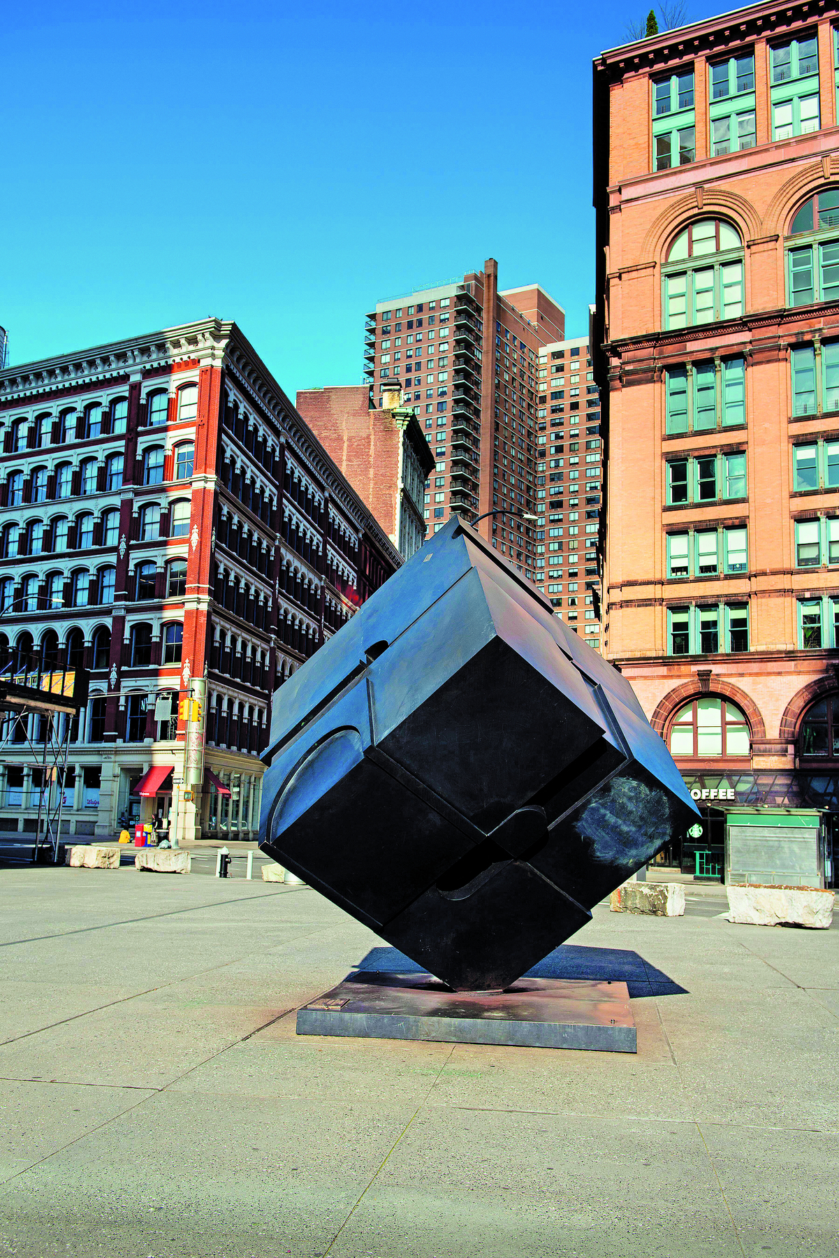 Fotografia. Escultura de aço no formato de cubo preto apoiado em um dos vértices em um patamar no chão. Ao fundo, prédios da cidade. O dia está ensolarado.