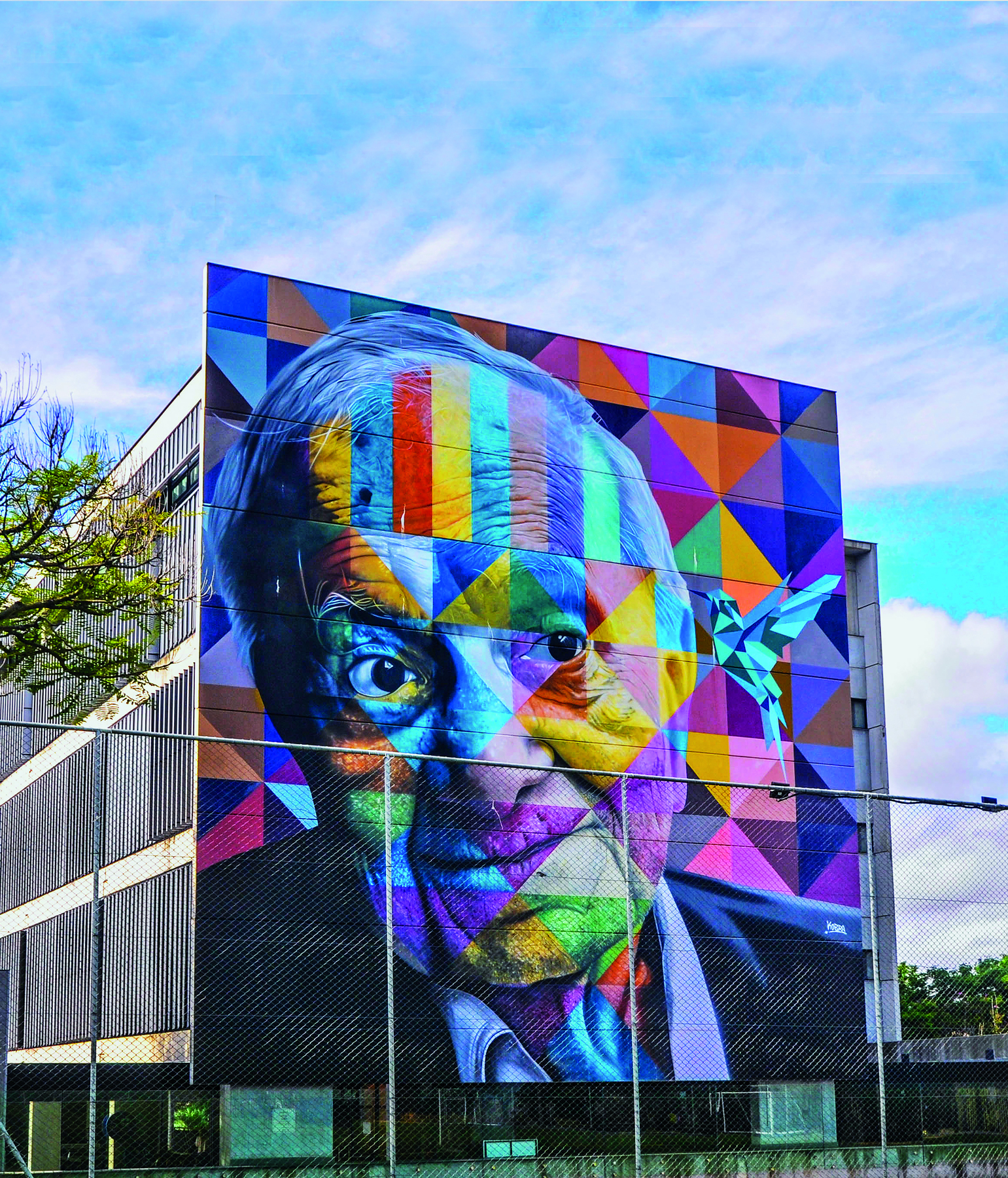 Foto. Fachada de um prédio com foto de Mario Quintana e grafite de Eduardo Cobra. Mario Quintana, senhor de cabelos grisalhos, está vestido com paletó. O grafite é colorido, pintado sobre a foto e toda a fachada, formado por quadriláteros de lados iguais, quadriláteros de lados opostos de mesma medida e triângulos. Há também no grafite um beija-flor azul, formado por figuras geométricas. Este toca seu bico na cabeça de Mario Quintana. O dia está ensolarado e com nuvens.