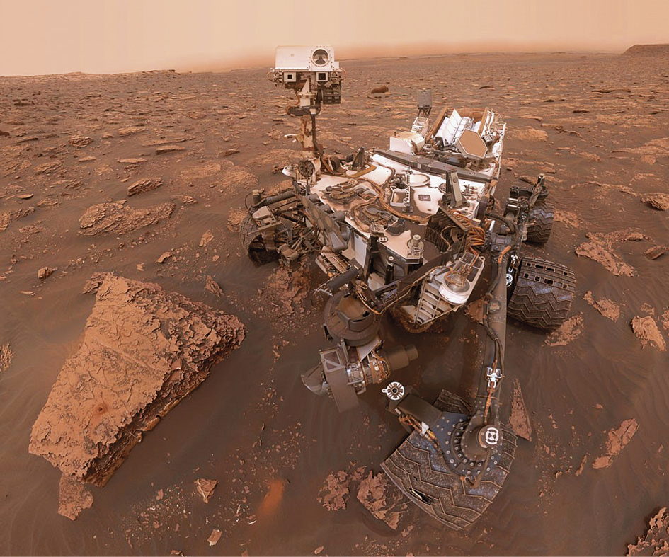 Fotografia. Vista do alto de superfície avermelhada com pedras e um jipe-robô com três rodas de cada lado sobre a superfície.