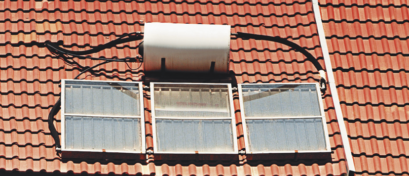 Fotografia. Vista do alto de um telhado com aparelho cilíndrico branco ligado por meio de fios pretos em três placas retangulares de vidro abaixo.