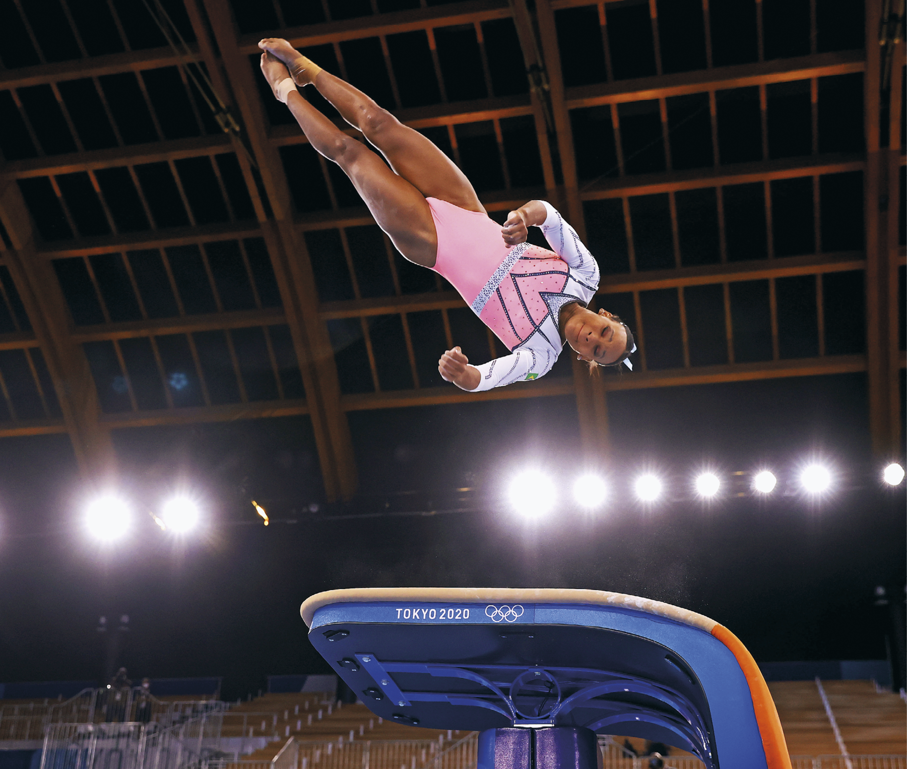 Fotografia. Mulher negra com os cabelos presos vestindo um colan rosa e branco com detalhes em prata, está saltando sobre um equipamento de ginastica olímpica. Ela está suspensa no ar de ponta cabeça. Ao fundo há luzes acesas. Abaixo há uma arquibancada.