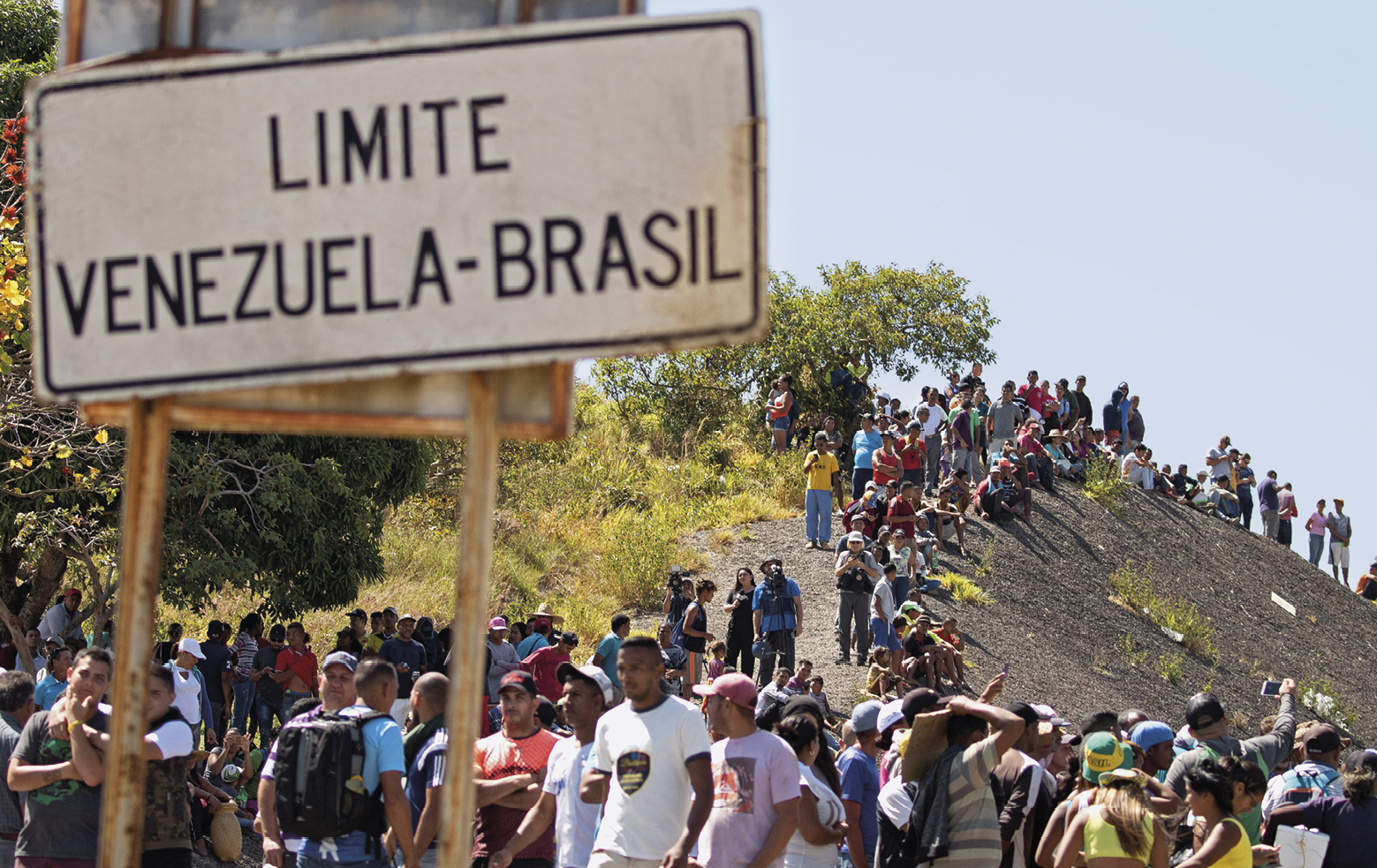 Foto. Aglomeração de pessoas ao pé de um morro de pedra, com vegetação verde. Há pessoas, subindo o morro, pessoas descendo o morro e pessoas paradas no pé do morro. Há uma placa com o escrito: "LIMITE VENEZUELA-BRASIL. O dia está ensolarado e sem nuvens.