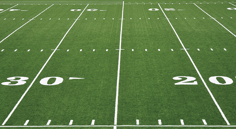 Fotografia. Vista parcial de um campo de futebol americano composto por linhas e traços. À esquerda, número 30 e, à direita, 20.