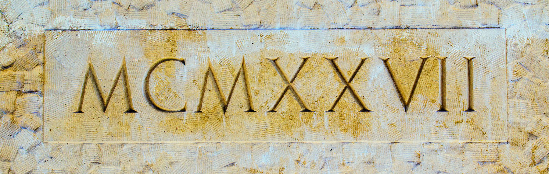 Fotografia. Letras MAIÚSCULAS  M C M X X V I I esculpidas em uma parede.