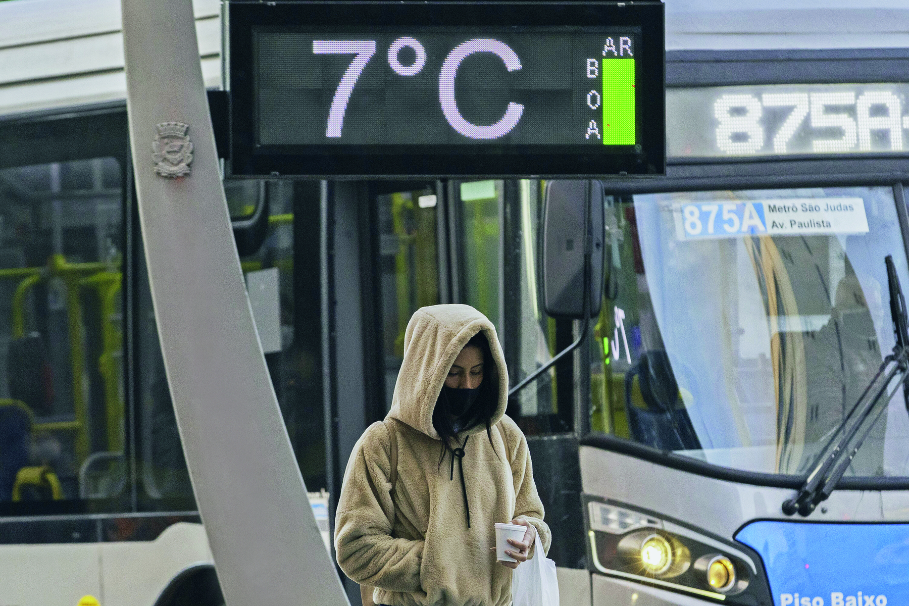 Fotografia. Uma pessoa caminhando na rua, vestida de blusa de frio bege, cachecol preto, com um copo de isopor e uma sacola nas mãos. Acima dela termômetro marcando 7 graus Celsius e umidade do ar, boa. Ao fundo, um ônibus circular. O dia está nublado.